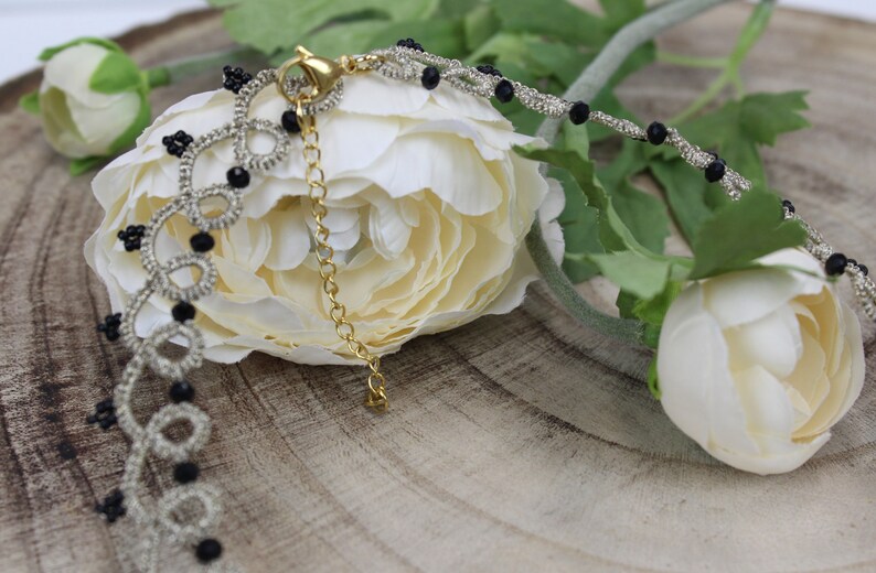 May include: A silver and black beaded necklace with a delicate chain and a clasp. The necklace is lying on a wooden surface with white flowers.