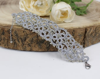 Puede incluir: Una pulsera plateada gris hecha a mano con un diseño delicado y de encaje. La pulsera presenta patrones intrincados y está adornada con pequeñas cuentas doradas. Tiene una cadena plateada para ajustar la talla y un cierre de broche. La pulsera se exhibe sobre una superficie de madera.