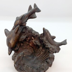 May include: A bronze sculpture of three dolphins leaping out of the water. The dolphins are arranged in a playful, dynamic pose, with one dolphin jumping over the other two. The sculpture is set on a base that resembles a rocky outcropping.