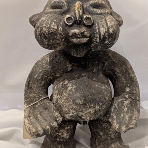 May include: A weathered, dark gray statue of a stout, humanoid figure with a large belly and bulbous cheeks. The statue has a simple, almost cartoonish face with a prominent nose and small eyes. The figure's arms are bent at the elbows, and its feet are wide and flat.