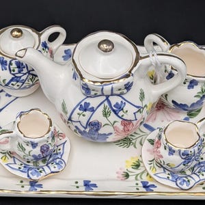May include: A miniature porcelain tea set with a white base, featuring blue floral patterns and gold trim. The set includes a teapot, creamer, sugar bowl, and two cups with saucers, all arranged on a matching rectangular tray.