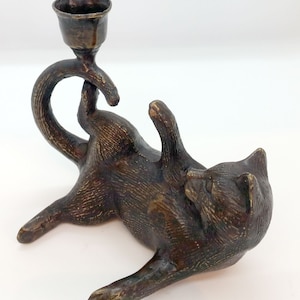 May include: Bronze cat-shaped candle holder. The cat is lying on its back with its paws in the air, supporting a candle cup. The candle holder has a textured surface and a dark bronze finish. A unique decorative item.