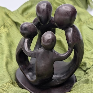 May include: A dark brown wooden sculpture depicting a family of four embracing. The figures are stylized with rounded heads and bodies, joined together in a circular embrace. The sculpture sits on a round base, against a green fabric background.