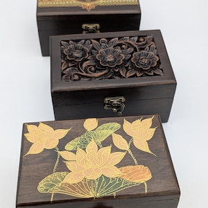 May include: Three decorative wooden boxes. The top box has a patterned lid with green, gold, and brown accents. The middle box features carved floral designs. The bottom box has a lotus flower design in gold and yellow.
