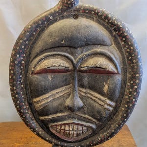 May include: A round, wooden tribal mask with a dark gray face and a snake-like design extending from the top. The mask features red accents around the eyes and mouth, with white and red dotted patterns around the perimeter.