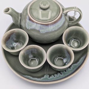 May include: A celadon green ceramic tea set, including a teapot with lid, four small cups, and a serving tray. The set has a crackle glaze finish with subtle color variations. The teapot has a curved spout and handle.