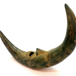 May include: A crescent-shaped, wooden sculpture with a face carved into it. The sculpture is painted in shades of green and brown, with a weathered appearance. The moon-shaped art piece has a hole in the center and is set against a white background.