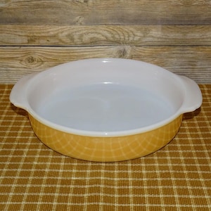 May include: An oval-shaped, vintage Pyrex baking dish. The interior is white, and the exterior features a yellow, grid-like pattern. It has two white handles on either side. The dish is sitting on a brown and tan checkered surface.