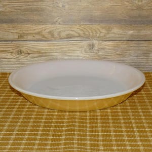 May include: A vintage, round pie dish with a white interior and a golden-yellow exterior. The dish has a slightly raised rim and is made of glass. The pie dish is sitting on a yellow and brown checkered surface.