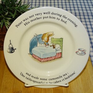 May include: A white ceramic plate with a picture of a bunny rabbit in a blue dress giving a cup of tea to another bunny rabbit in bed. The plate has the text "Peter was not very well during the evening. His mother put him to bed, and made some camomile tea; One table-spoonful to be taken at bed-time."