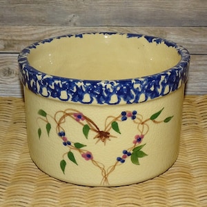 May include: A cream-colored ceramic bowl with a blue and white speckled rim. The bowl has a hand-painted design of a heart made of vines with green leaves and pink and blue berries.