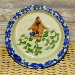 May include: A ceramic pie plate with a blue and white rim. The center of the plate features a hand-painted design of a birdhouse with a bluebird perched on the edge, surrounded by green ivy.
