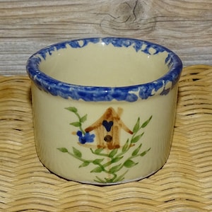 May include: A small, cream-colored ceramic pot with a blue rim and a hand-painted design of a birdhouse with bluebirds and green leaves.