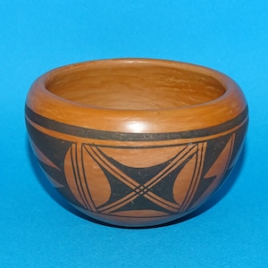 May include: A handcrafted, round, earthenware bowl with a reddish-brown exterior. The bowl features a black and brown geometric pattern around the exterior. The interior is a lighter shade of brown. The bowl is set against a blue background.