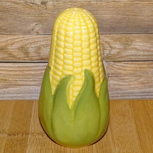 May include: A ceramic salt shaker shaped like an ear of corn. The shaker is yellow with a textured surface to mimic corn kernels, and green leaves form the base. The design is a novelty kitchen item.