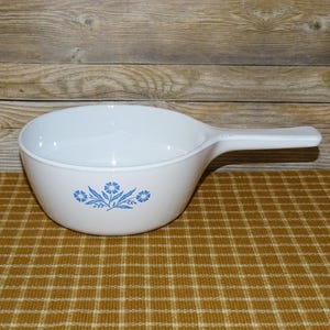 May include: A white ceramic saucepan with a single handle and a blue floral pattern on the front. The bowl is round with a smooth interior and a flat base, featuring a stylized blue flower design centered on the exterior side of the vessel.