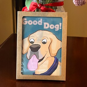 May include: A framed print featuring a cartoon illustration of a golden Labrador Retriever with its tongue sticking out. The text "Good Dog!" is printed above the dog. The frame is made of brown cardboard.