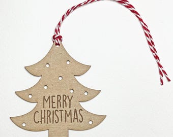 May include: A wooden Christmas tree ornament with the words "MERRY CHRISTMAS" engraved on it. The tree is light brown with small circular cutouts and is suspended by a red and white twine. The ornament is a festive holiday decoration.