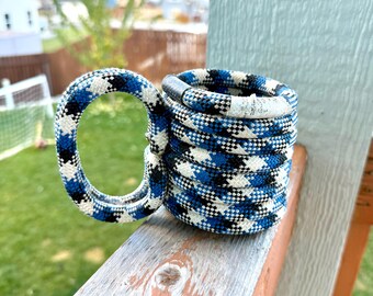 May include: A handmade mug wrapped in a blue, black, and white patterned rope. The mug has a rounded handle and a cylindrical body. The mug is sitting on a wooden surface with a green lawn and houses in the background.