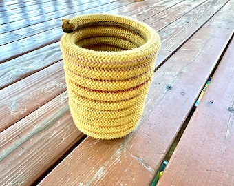 May include: A yellow rope basket with a cylindrical shape. The basket has a woven texture with horizontal red stripes. The basket is sitting on a wooden deck.