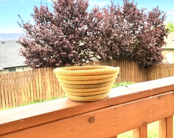 May include: A woven wicker basket with a natural tan color. The basket is sitting on a wooden railing with a green grassy area in the background.