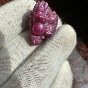 May include: A carved, deep red gemstone dragon figurine. The dragon is holding a sphere and is detailed with scales and facial features. The figurine is held in a white glove.