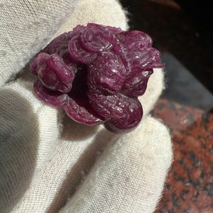 May include: A detailed, carved figurine of a frog in a deep burgundy color. The frog is depicted in a seated position with intricate details, held in a white glove. The sculpture has a smooth, polished finish, and the frog's features are clearly defined.