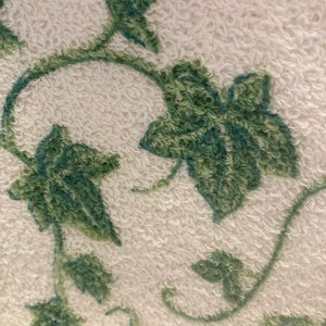 May include: Close-up of a white terry cloth towel with a green vine and leaf pattern. The leaves and vines are a darker shade of green, creating a textured, three-dimensional effect. The towel's surface has a soft, looped texture.