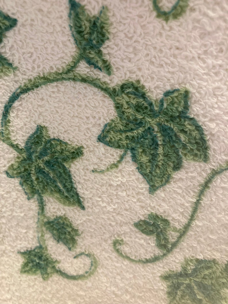 May include: Close-up of a white terry cloth towel with a green vine and leaf pattern. The leaves and vines are a darker shade of green, creating a textured, three-dimensional effect. The towel's surface has a soft, looped texture.