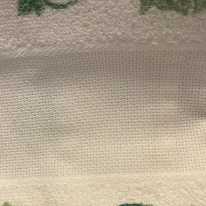 May include: Close-up of a white towel with a textured weave. The towel has a folded section, revealing a cross-stitch pattern. Green floral designs are visible at the bottom edge of the towel. The background is a wooden surface.