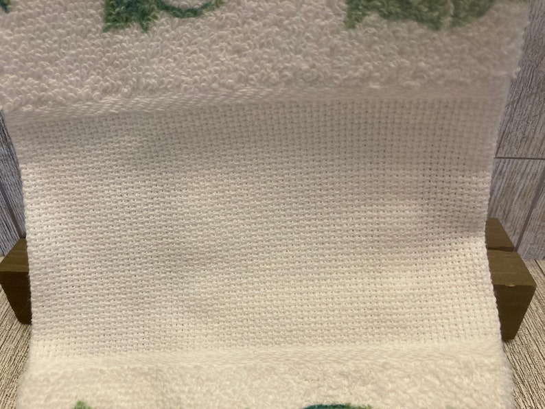 May include: Close-up of a white towel with a textured weave. The towel has a folded section, revealing a cross-stitch pattern. Green floral designs are visible at the bottom edge of the towel. The background is a wooden surface.