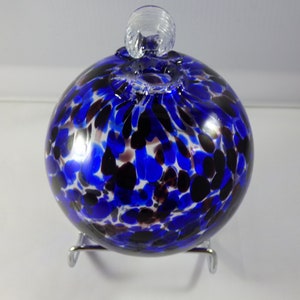 May include: A round, blue glass ornament with a speckled pattern of black and dark red spots. The ornament has a clear glass loop at the top and sits on a silver metal stand. The glass sphere is approximately 4 inches in diameter.
