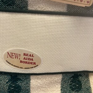 May include: Close-up of a white Aida cloth with a black border, and a green and white plaid pattern. An oval sticker reads "NEW! REAL AIDA BORDER" with text that says "EASIER TO STITCH AND...HIDES THE BACK!"