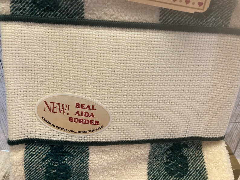 May include: Close-up of a white Aida cloth with a black border, and a green and white plaid pattern. An oval sticker reads "NEW! REAL AIDA BORDER" with text that says "EASIER TO STITCH AND...HIDES THE BACK!"