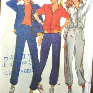 May include: A sewing pattern illustration featuring three athletic outfits. One includes a blue zip-up jacket and matching pants. Another shows a red short-sleeved zip-up jacket with blue pants. The third displays a light gray zip-up jacket and pants.