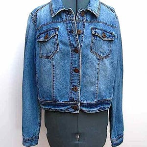 May include: A light blue denim jacket with a classic design. The jacket features two chest pockets, brown buttons, and a collared neckline. The garment is displayed on a mannequin, showcasing its structure and fit. A timeless piece for casual wear.