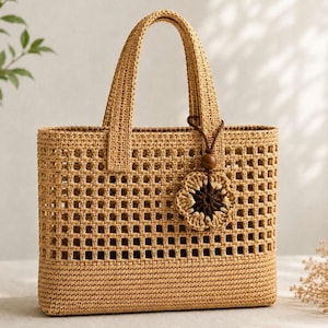 May include: A woven tote bag in a light tan color, featuring a rectangular shape with a square grid pattern. The bag has two handles and a decorative flower charm. The bag is made of a natural fiber material.