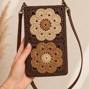 May include: A rectangular crochet crossbody bag in dark brown featuring two large floral motifs. The top flower is cream with a brown center, and the bottom flower is light brown with a cream center. It includes a thin brown leather-like strap.
