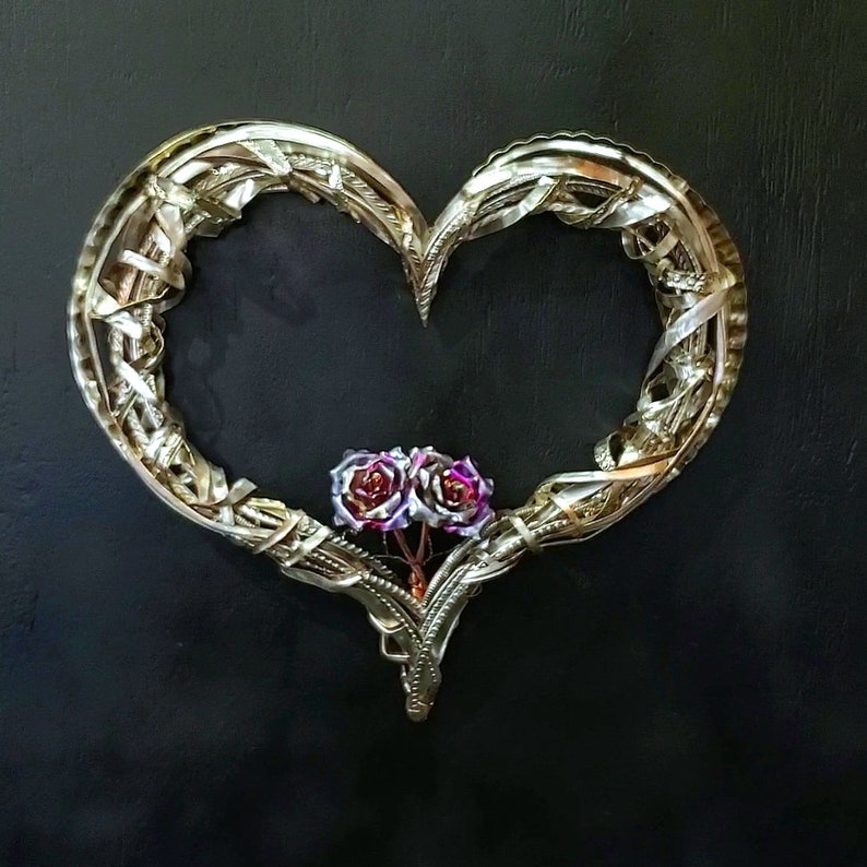 May include: A metal heart-shaped wreath with a delicate, interwoven design. Two small, pink and purple roses are nestled in the center of the wreath.