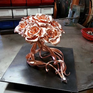 May include: A metallic sculpture of a bouquet of roses crafted from polished copper. The roses feature layered, curved petals with a reflective finish. Below the flowers, several long, thin copper ribbons and wires extend outward, terminating in a small, dark, faceted metal point.