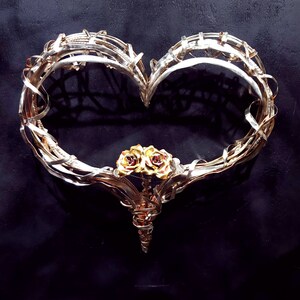 May include: A heart-shaped metal sculpture made of intertwined silver and gold wire. The sculpture features two golden roses at the bottom.