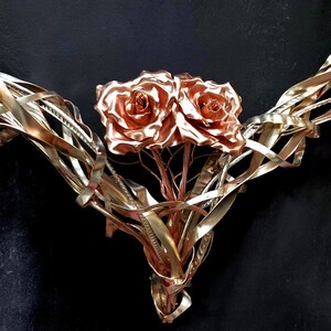May include: A metal sculpture of two roses with intertwined stems, crafted from copper wire. The roses are a warm copper color and the stems are a lighter gold color.
