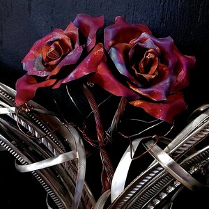 May include: Two copper roses with a purple and red iridescent finish are intertwined with silver wire. The roses are arranged in a bouquet and are set against a dark background.