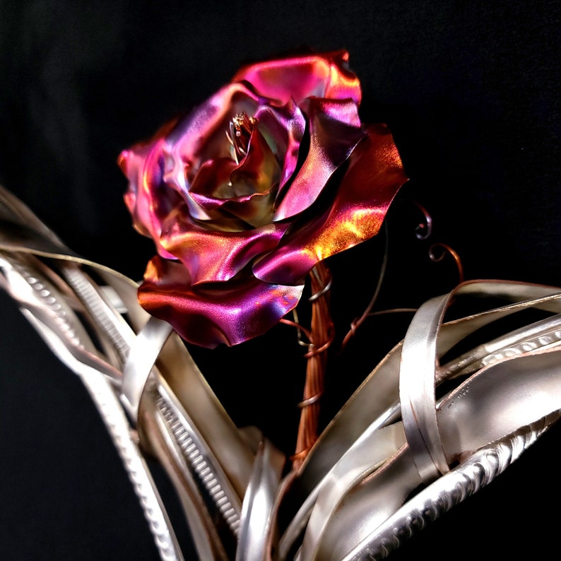 May include: A metal rose sculpture with a pink and orange iridescent finish. The rose is set in a silver metal base with a twisted, vine-like design.