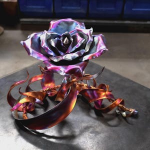 May include: A metal rose sculpture featuring iridescent blue, purple, and magenta petals with a central point. The base consists of curved, ribbon-like metal strips in shades of copper, bronze, and violet, creating a dynamic, flowing appearance.
