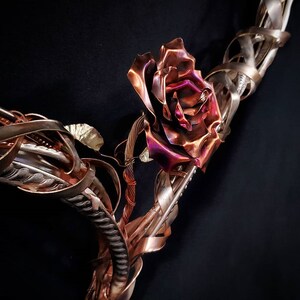 May include: A metal sculpture of a rose with a copper and silver finish. The rose is made of twisted metal and has a textured surface. The sculpture is a unique and artistic piece.