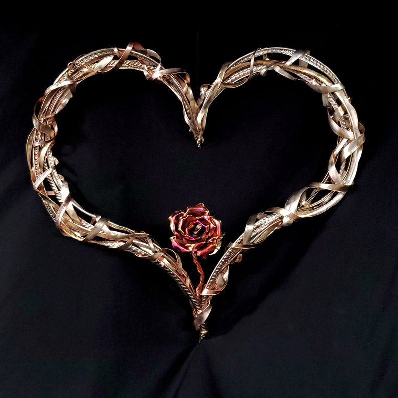 May include: A heart-shaped metal sculpture with a rose design. The heart is made of intertwined metal wires, creating a delicate and intricate pattern. The rose is a single bloom with a pink and red color gradient.