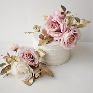 May include: Two arrangements of artificial flowers, one on top of a white cake and one on a white surface. Both arrangements feature pink and white roses with gold-painted leaves.