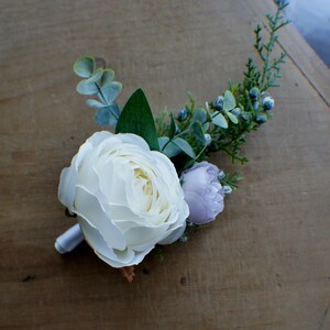 May include: A white rose with a light purple flower and green foliage, arranged in a small bouquet.