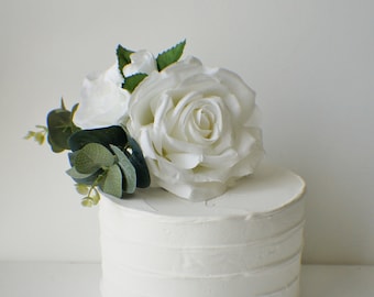 Op de afbeelding: Een witte cake met witte frosting en een witte roos met groene bladeren erop.
